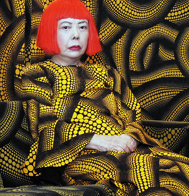 Portrait of artist Yayoi Kusama. She is wearing a short red wig along with a yellow and black patterned dress filled with dots.
