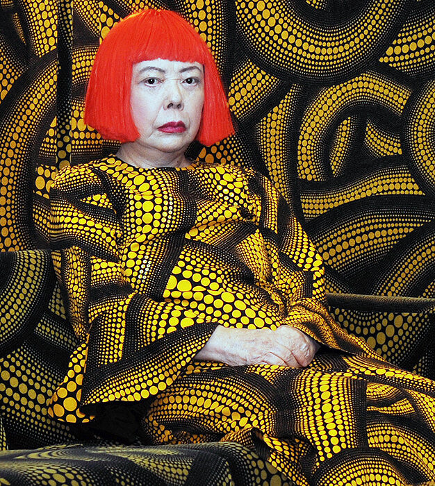 Portrait of artist Yayoi Kusama. She is wearing a short red wig along with a yellow and black patterned dress filled with dots.