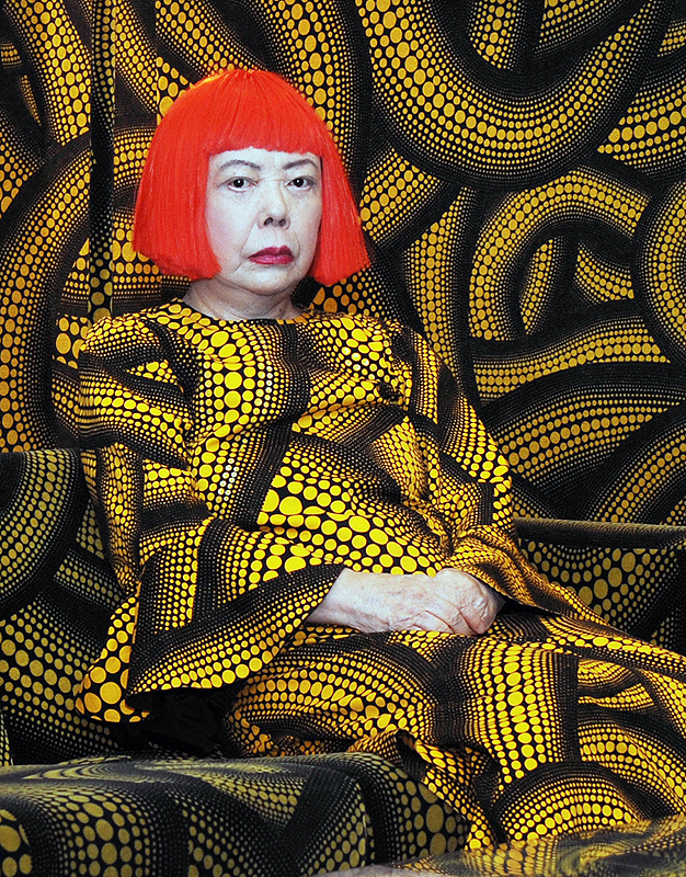 Yayoi_Kusama_Feature Portrait of artist Yayoi Kusama. She is wearing a short red wig along with a yellow and black patterned dress filled with dots.